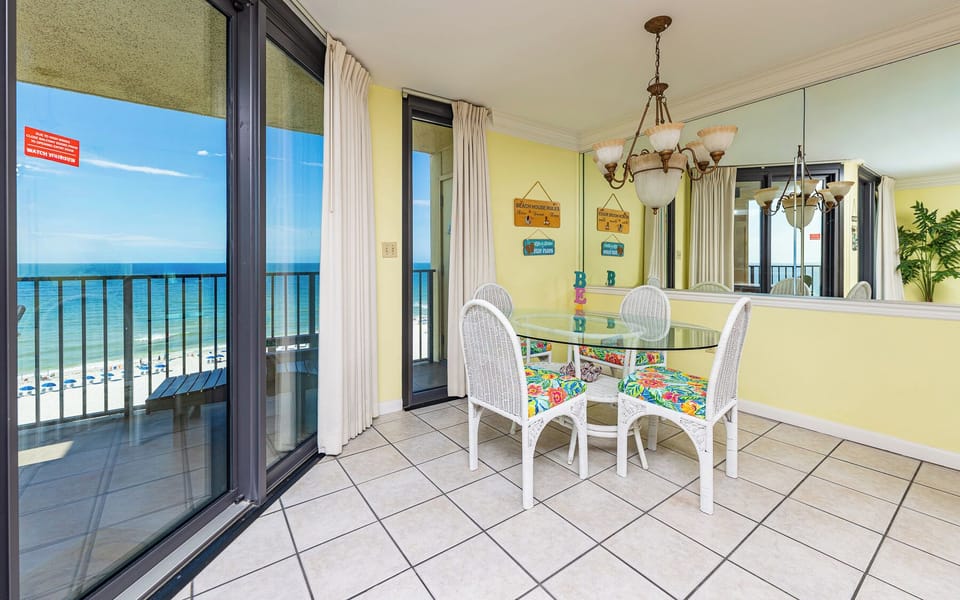 Dining area with a glass table, colorful cushioned chairs, and sliding doors leading to a balcony with a beach front view