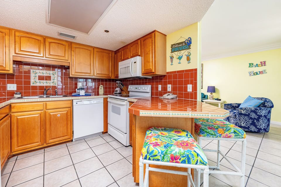 Kitchen with tiled breakfast bar, featuring wooden cabinetry and a cozy living area visible in the background