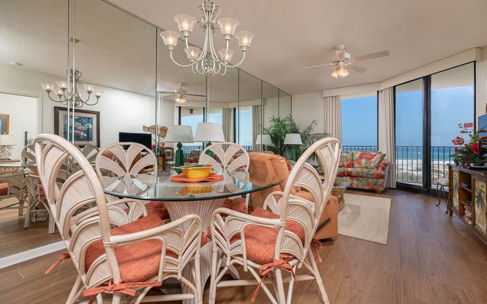 Dining area features a glass table surrounded by rattan chairs, with a vibrant view of the beach through large windows