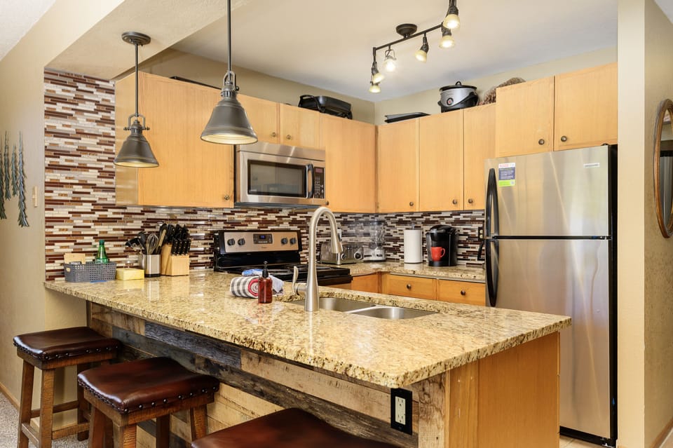 Updated kitchen with granite counter tops, wooden cabinetry, stainless steel appliances, and a tiled backsplash, with pendant lighting.