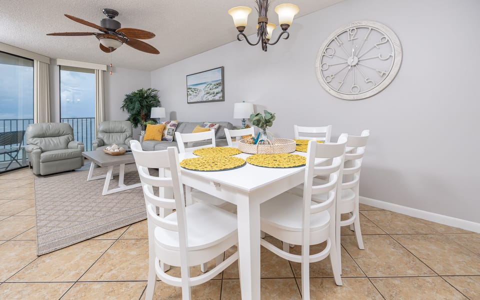 Dining area features a white table with yellow placemats, surrounded by chairs, and a cozy living space with beach views