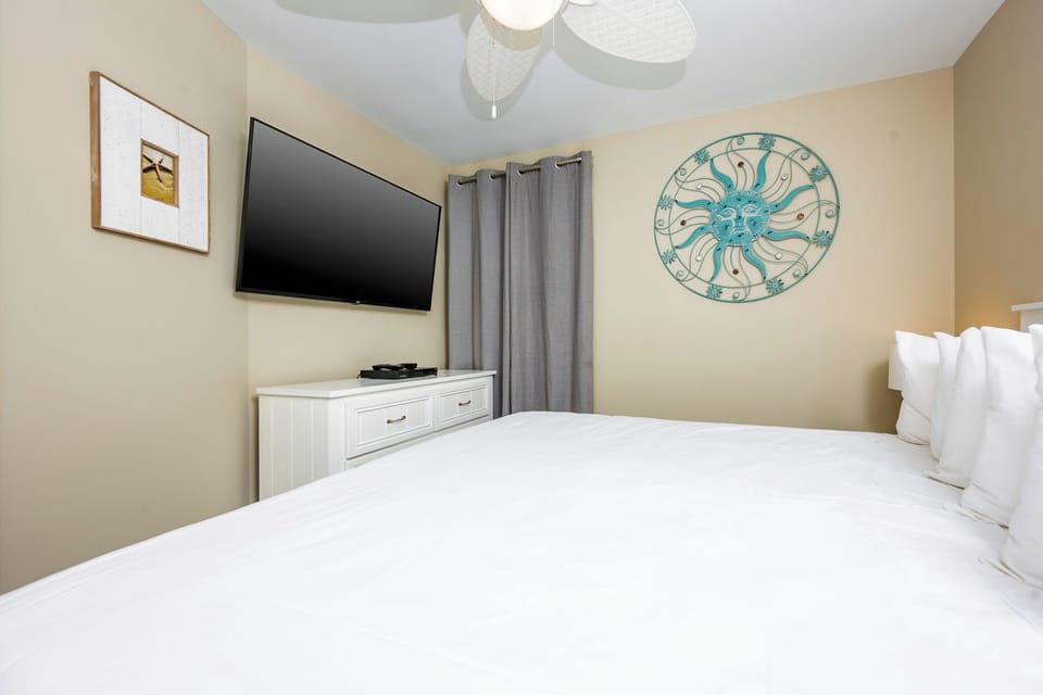 This cozy bedroom features a large TV, a stylish sun-themed wall decor, and soft white bedding for a restful stay