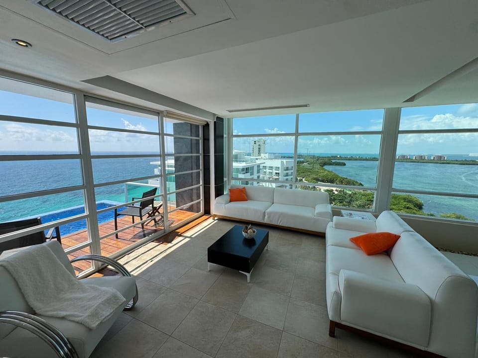 Corner seating area with white sofas, orange pillows, and glass walls revealing ocean and lagoon views.