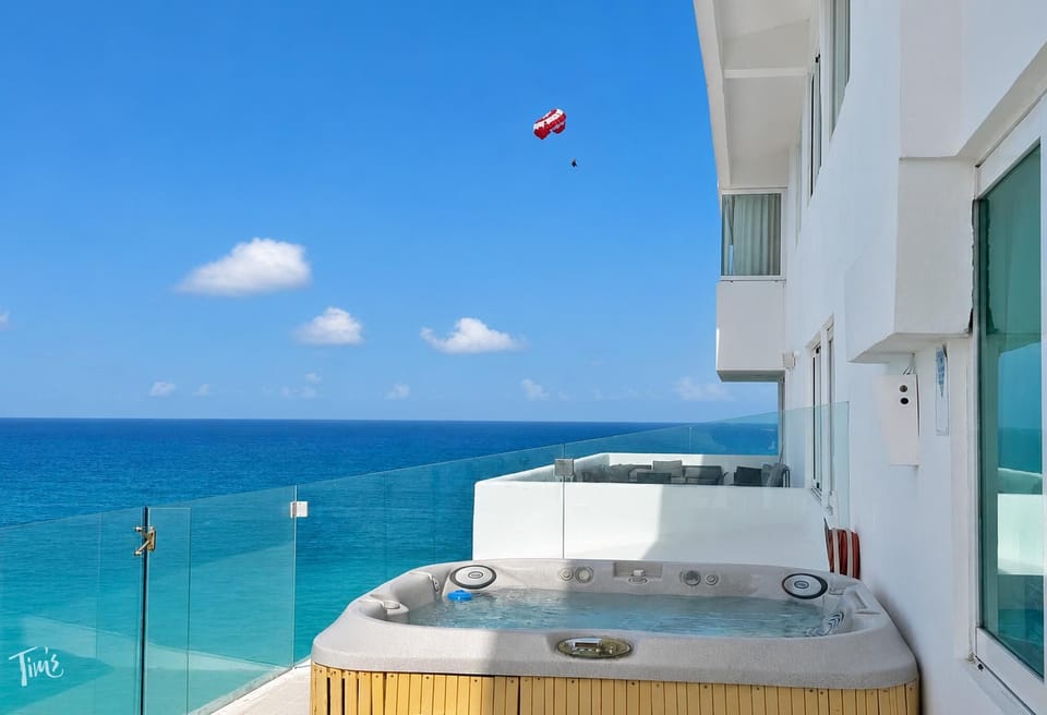 Corner hot tub on the balcony with glass railing overlooks turquoise ocean and a parasailer in the bright blue sky.