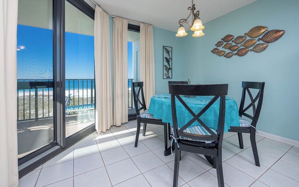 Bright dining area features a teal table with black chairs, offering a view of the beach through expansive glass doors