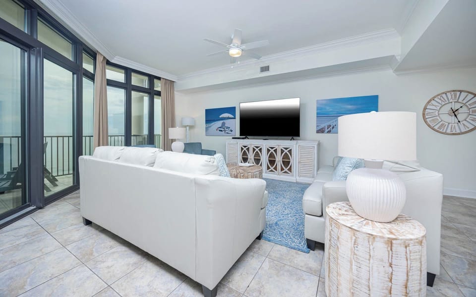 This living area features a large TV, light-colored furniture, and stunning beachfront views through expansive glass windows