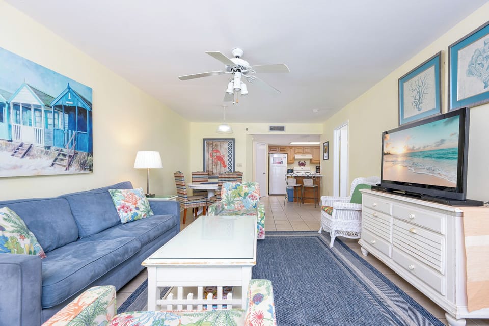 Living area featuring a blue sofa with queen sleeper, colorful chairs, and large TV, leading to a kitchen in the background