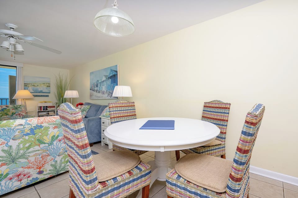 A round dining table with colorful woven chairs complements the cozy living area, featuring beach-themed artwork and lamps