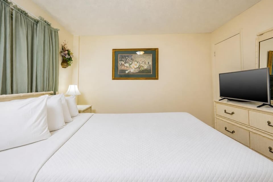 The bedroom features a bed with white linens, a TV, and soft green curtains, creating a serene atmosphere for relaxation
