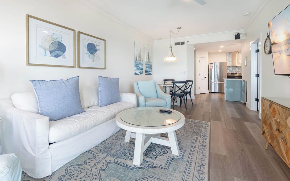Relax in this sleek living space with cozy seating, a glass-top table, and coastal art to enhance your beach getaway
