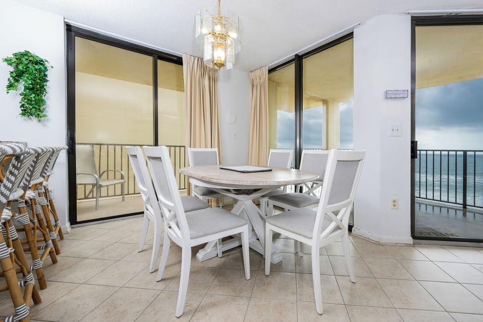 Bright dining area featuring a round table with six chairs, accented by a stylish chandelier and views of the beachfront