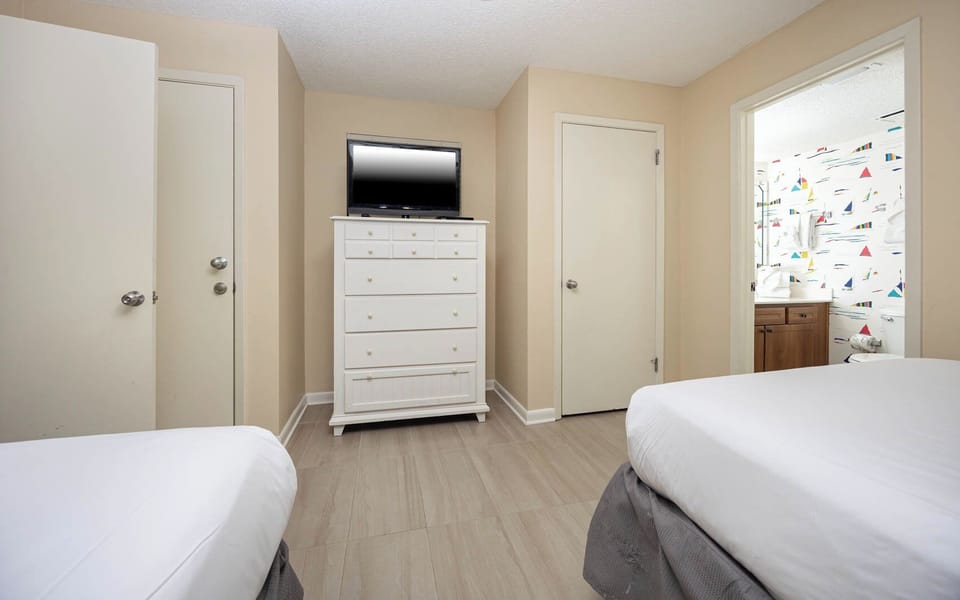 Bright guest bedroom featuring two double beds, a white dresser, and a TV, with colorful nautical decor in the bathroom