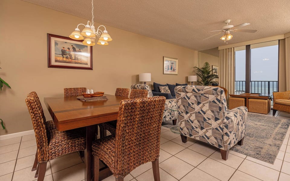 Dining table with woven chairs complements the cozy living area, featuring patterned armchairs and a view of the beach beyond