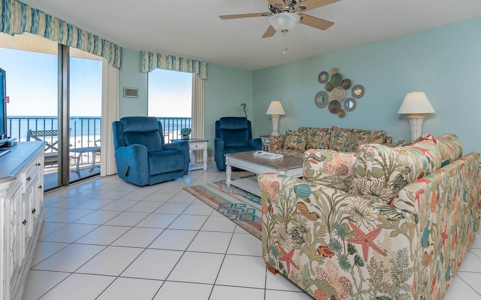 The living room features vibrant floral-patterned sofas, cozy blue recliners, and a view of the beach