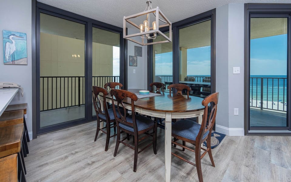 The dining area features a wooden table surrounded by six chairs, with expansive views of the beach through large windows