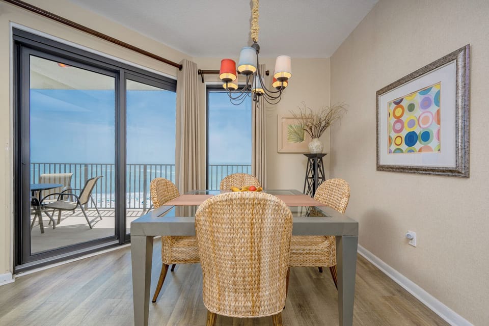 Dining area features a glass-top table surrounded by woven chairs, with sliding doors revealing a beachfront view