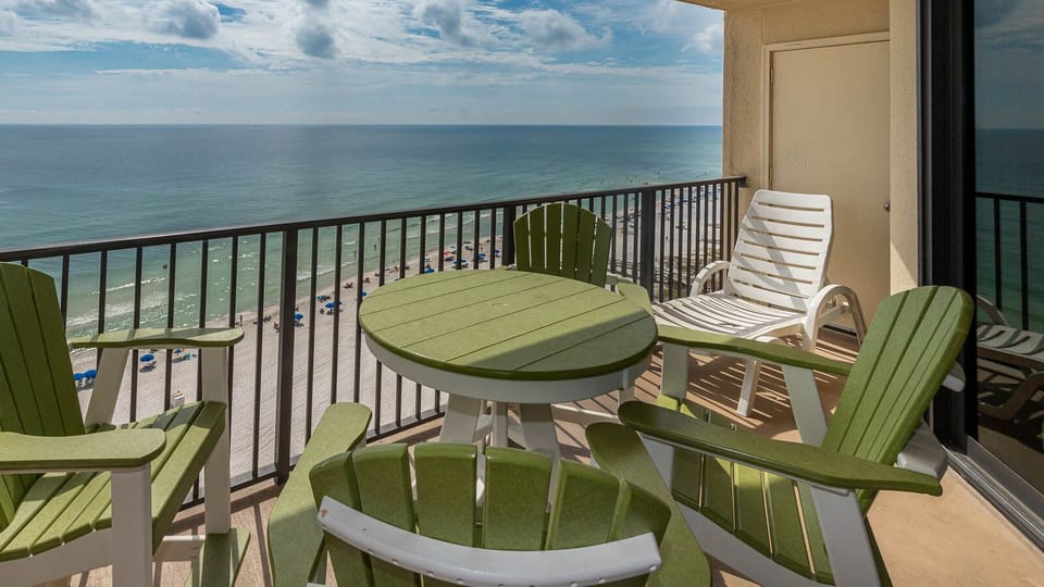 The balcony features green and white outdoor seating with a view of the beach and umbrellas lining the shore below