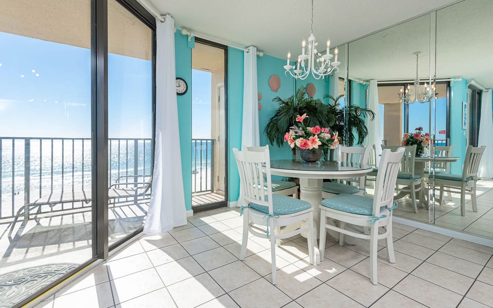 Bright dining area with a round table, white chairs, and vibrant floral centerpiece, offering a view of the beach