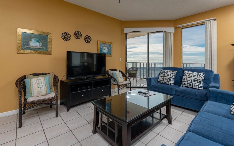 The living room features a blue sofa with patterned cushions and queen sleeper, a flat-screen TV, and a view of the beach