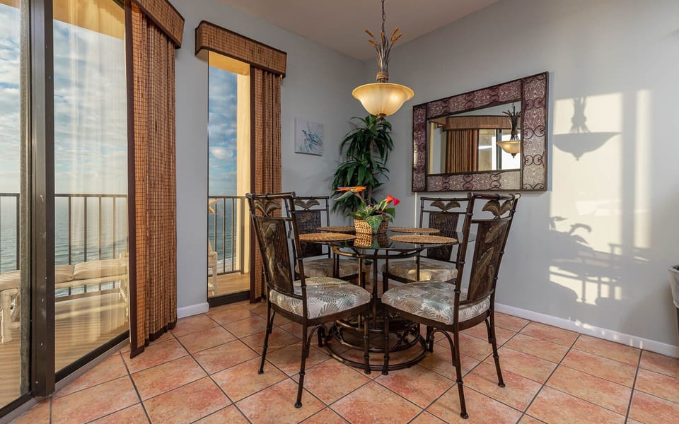 A round dining table with tropical chairs sits near large windows, offering a view of the beach and bright natural light