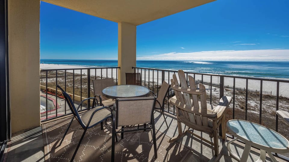 The balcony features a glass-top table with four chairs, offering a panoramic view of the tranquil beachfront and blue sky