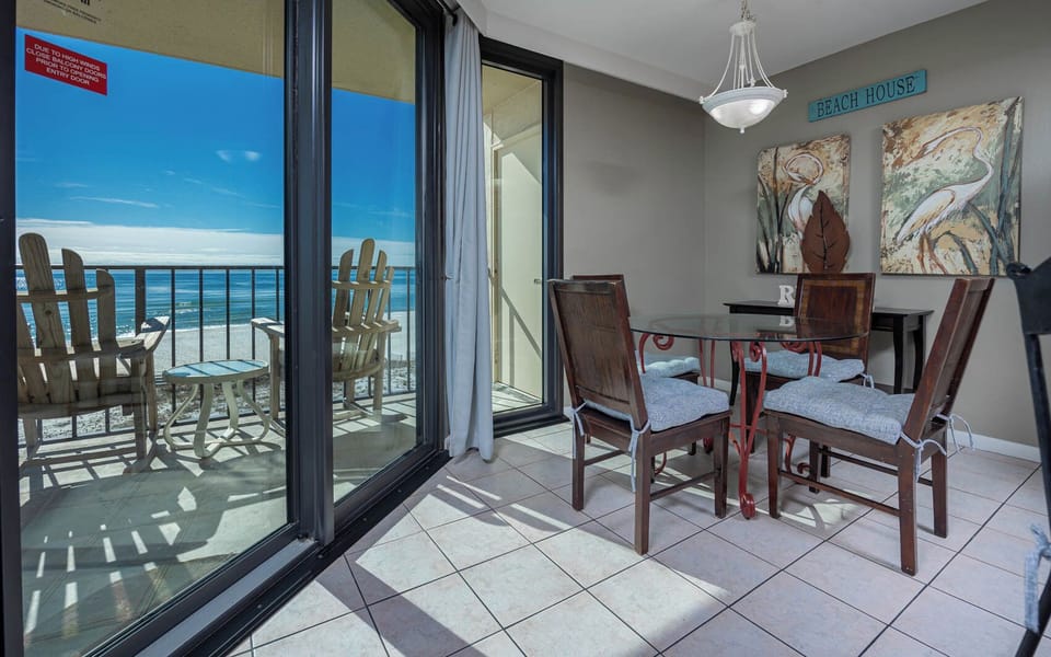 The dining area features a glass table with wooden chairs, offering a view of the beach through large sliding doors