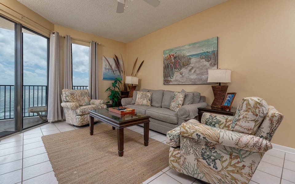 Cozy living area features a gray sofa with queen sleeper, two patterned chairs, and large balcony with a view of the beach
