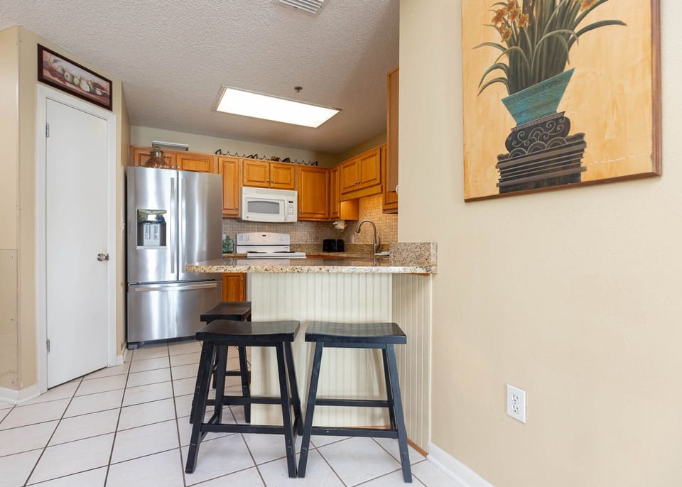 The kitchen features warm wood cabinetry, a granite countertop breakfast bar with two black stools, and a large refrigerator