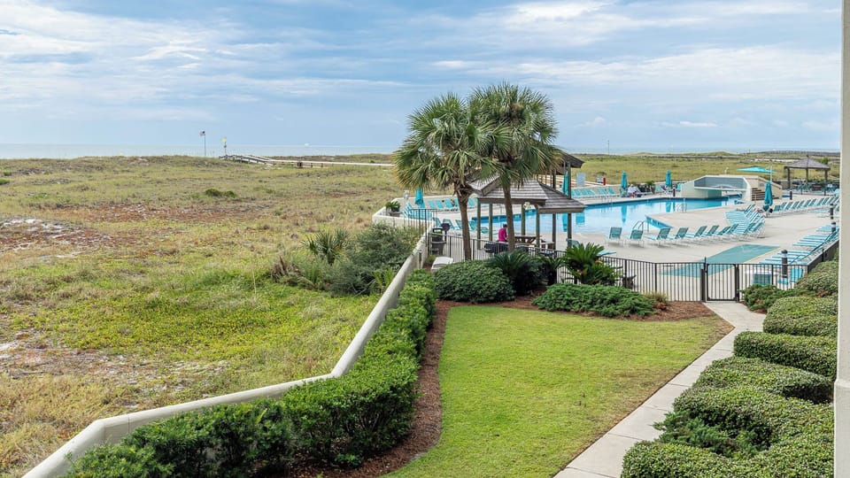 Lush green landscaping frames the inviting pool area, with lounge chairs and a view of the beach in the distance