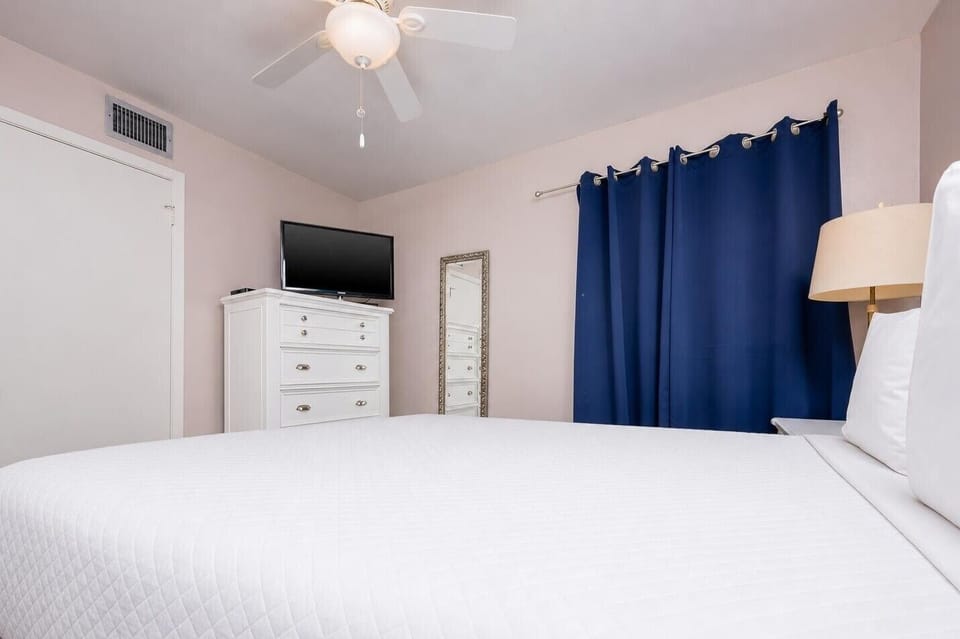 This guest bedroom features a white queen bed, a flat-screen TV, and rich blue curtains for a relaxing atmosphere