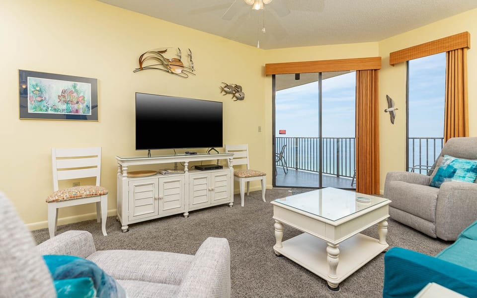 Living area features a large TV, comfortable seating, and sliding glass doors opening to a balcony with beach front views