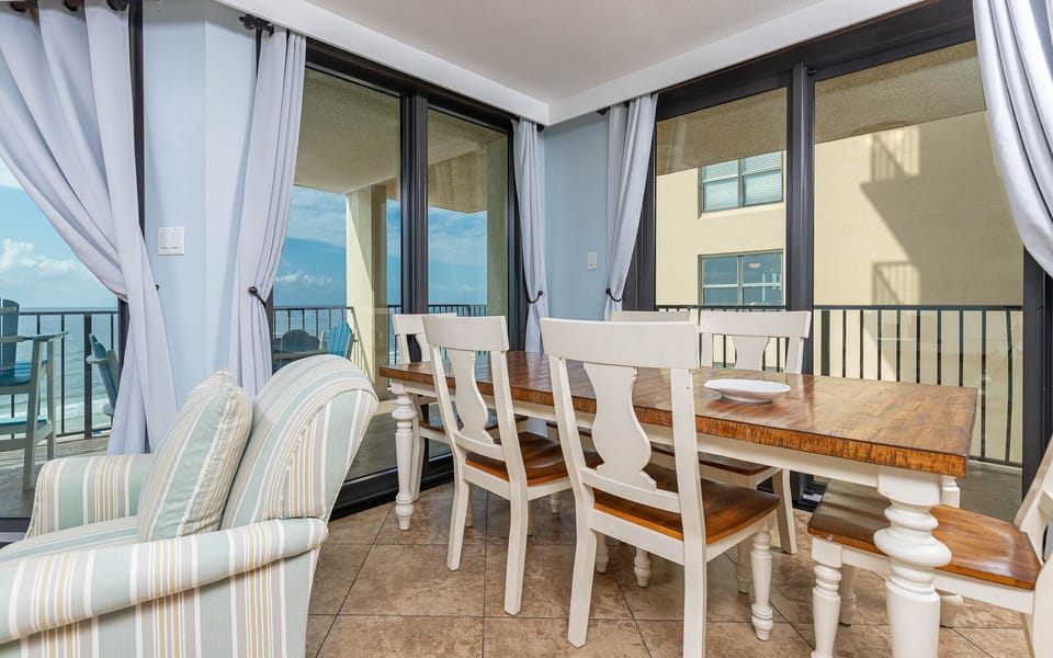 A light-filled dining area features a rustic wooden table surrounded by white chairs, with beach views