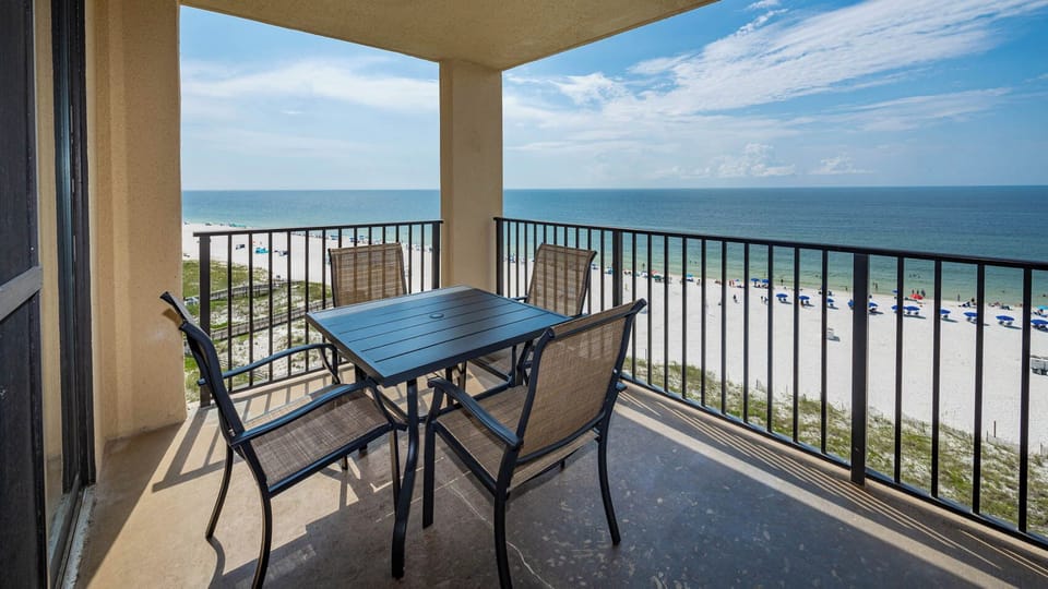 The balcony features a dining table with four chairs, offering a beautiful view of the beach and tranquil waters