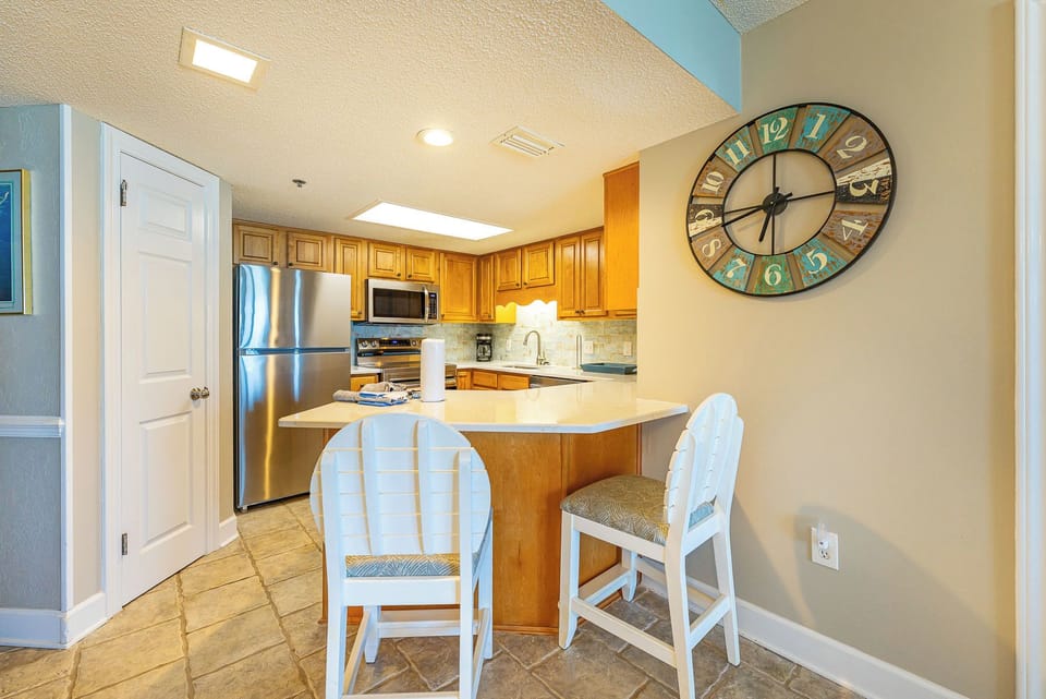 Bright wood cabinetry complements the kitchen, featuring a breakfast bar with white stools and a stylish wall clock