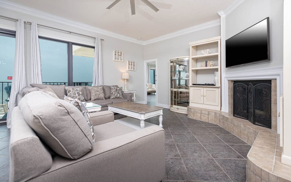 The living area features a gray sofa with queen sleeper, fireplace, flat-screen TV, and a lovely beach view from the balcony