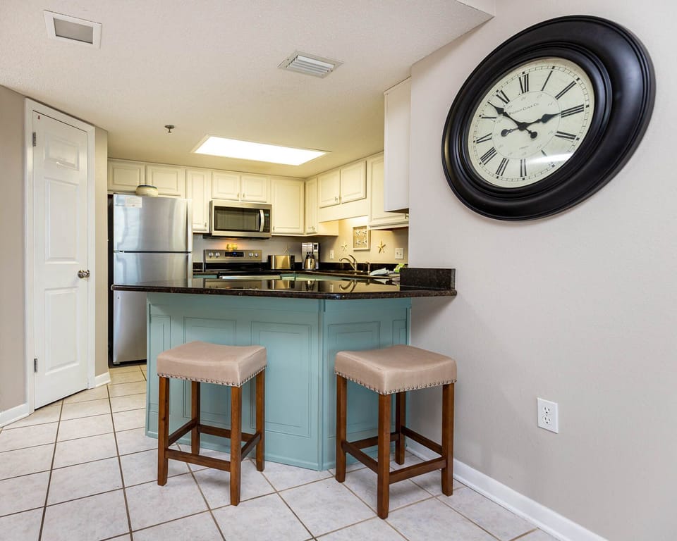 Bright kitchen area features a sleek island with bar stools, stainless appliances, and a stylish clock on the wall