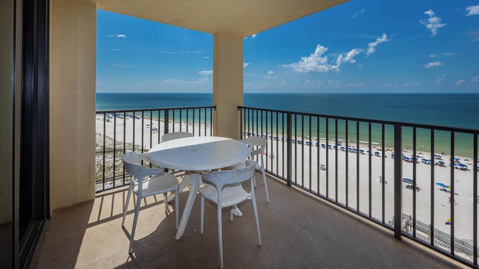 A round white table with four chairs overlooks a beach front, showcasing white sand and umbrellas under a clear blue sky