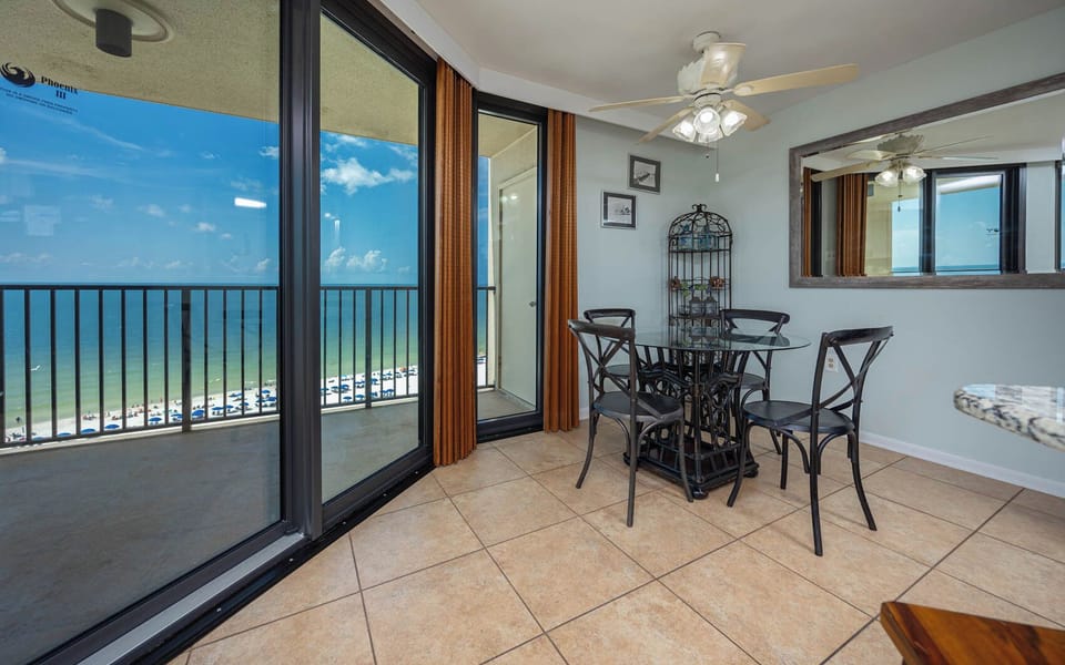 The dining area features a glass table with black chairs and offers a panoramic view of the beach through large sliding doors