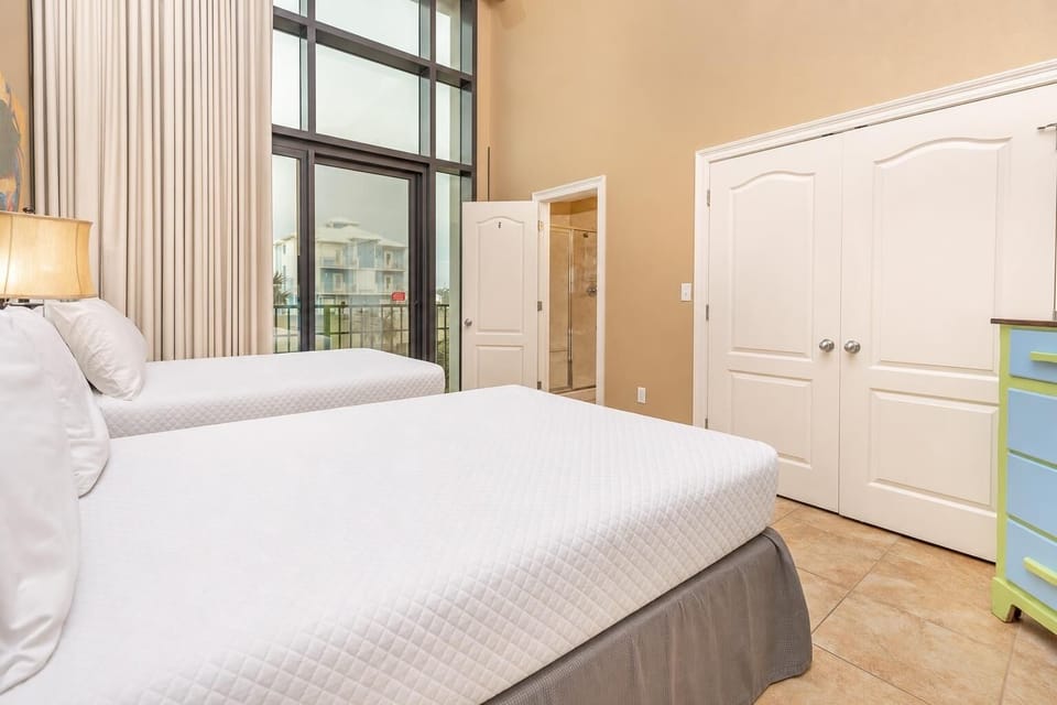 This bright bedroom features two full beds, a colorful dresser, and a TV, with large windows offering a view of the outdoors