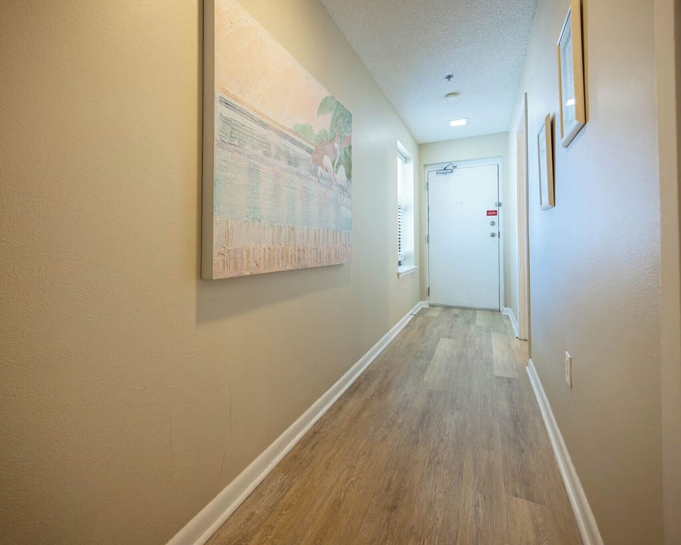 Welcoming hallway features light wood flooring, soft colors, and artwork, leading to a door and natural light from a window
