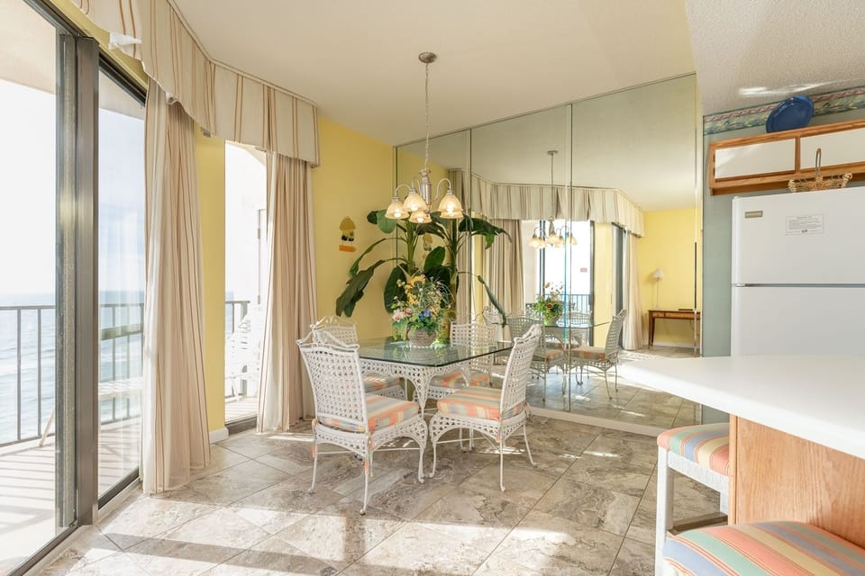 Bright dining area features a glass table with colorful chairs, surrounded by a view of the beach