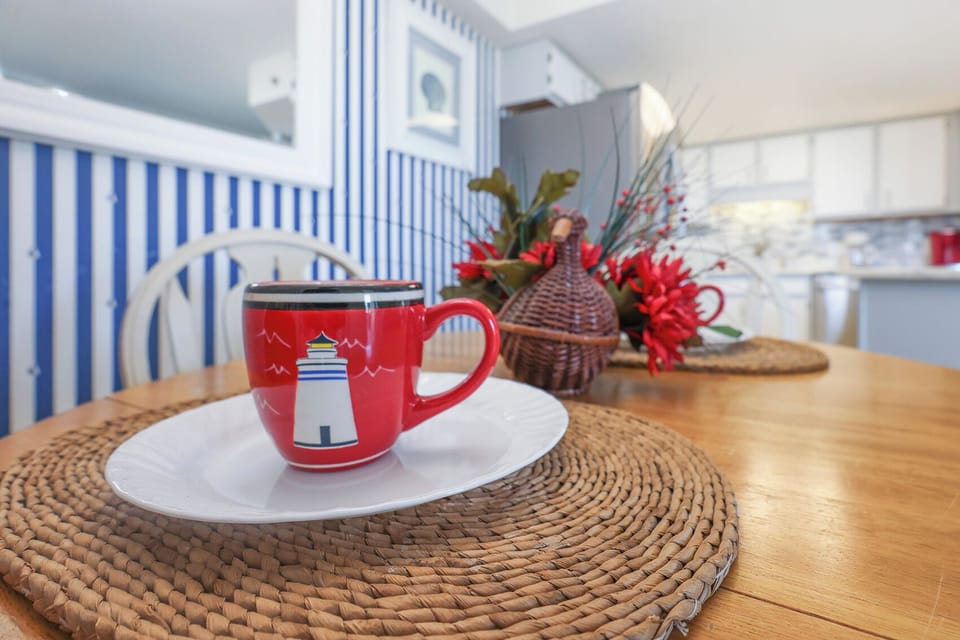 A colorful lighthouse mug sits on a white plate atop a woven placemat, complemented by vibrant floral decor