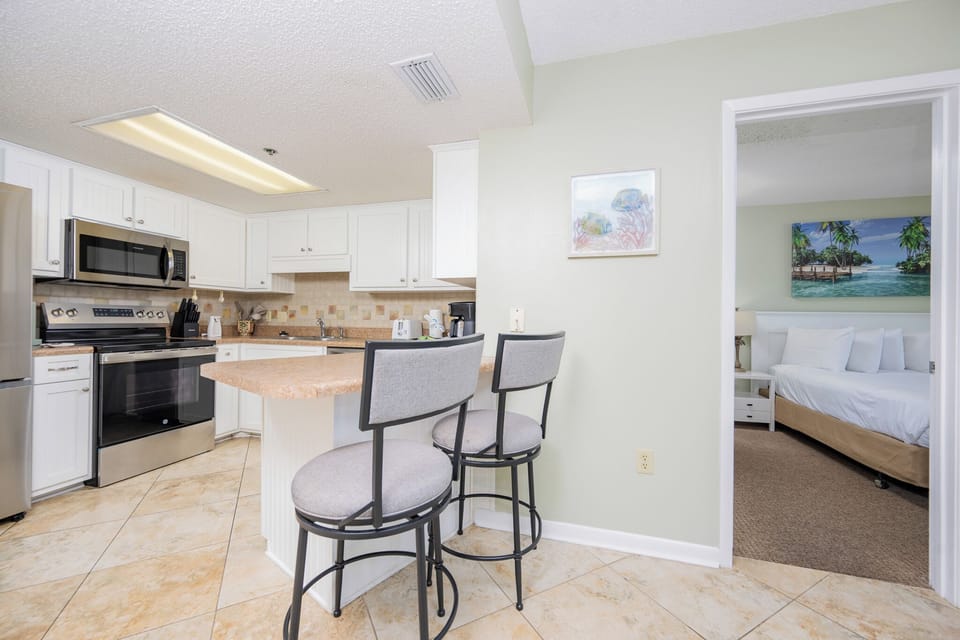 Bright kitchen with a breakfast bar, featuring white cabinetry, stainless appliances, and a view into a cozy guest bedroom