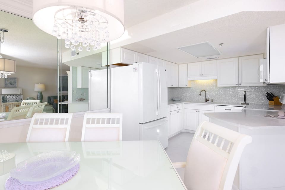 White cabinetry and a sleek kitchen complement the dining area, featuring a glass table and elegant chandelier overhead