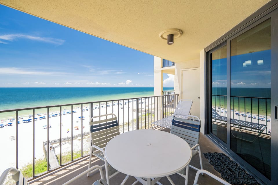 The balcony features a round table and chairs, offering a clear view of the beach and vibrant blue waters below