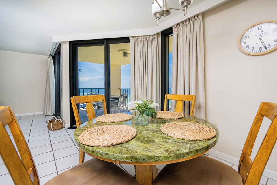 A round green marbled dining table with woven placemats is set near large glass doors leading to the beachfront balcony