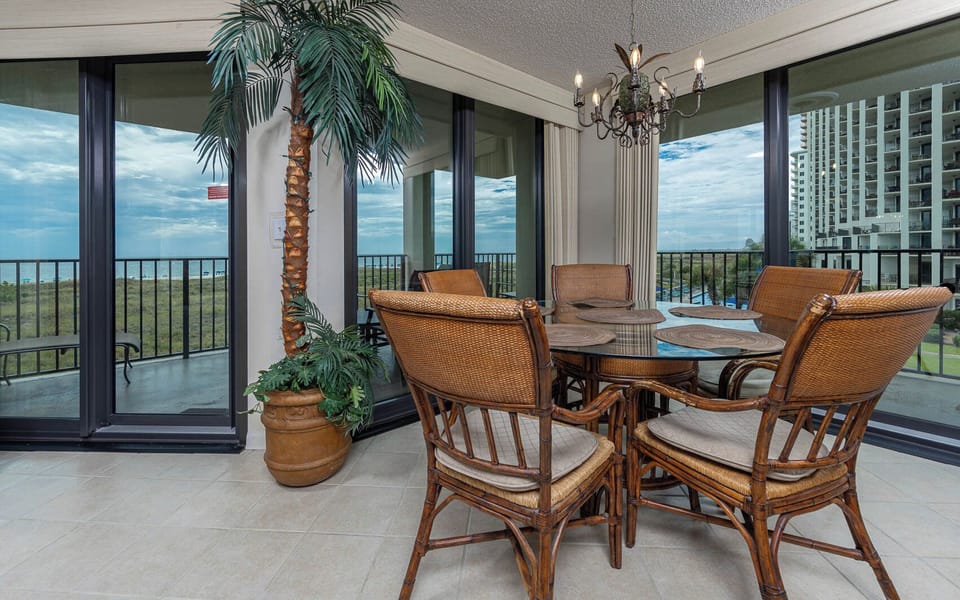 Dining area featuring a glass table surrounded by wicker chairs, with a view of the beach visible through large windows