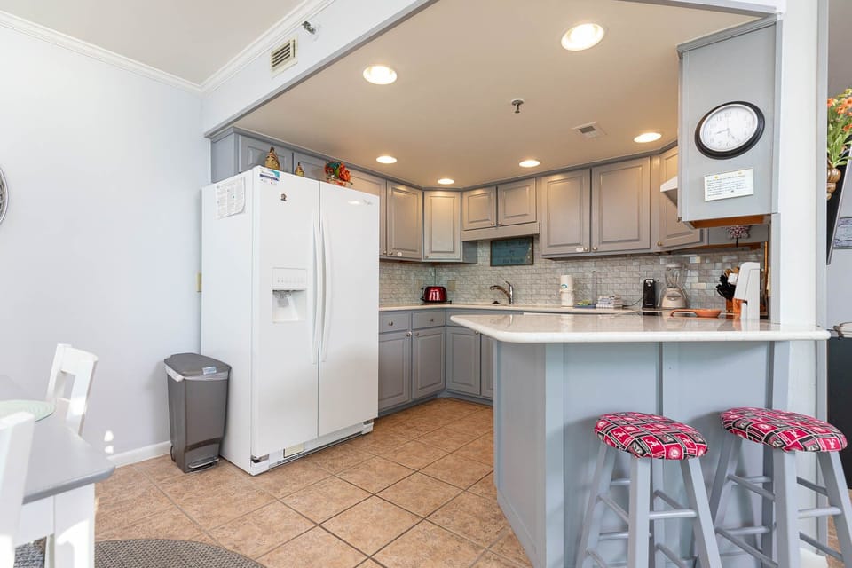 The well-equipped kitchen features light gray cabinetry, white appliances, and colorful bar stools at the counter