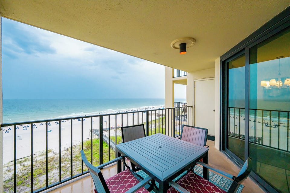 The balcony features a table and chairs with a view of the beach and distant umbrellas against a blue sky