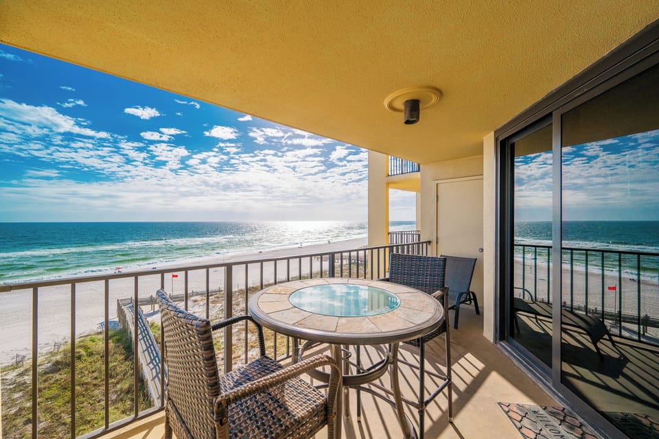 The balcony features a round table and chairs, offering a panoramic view of the sparkling beachfront and vibrant blue sky
